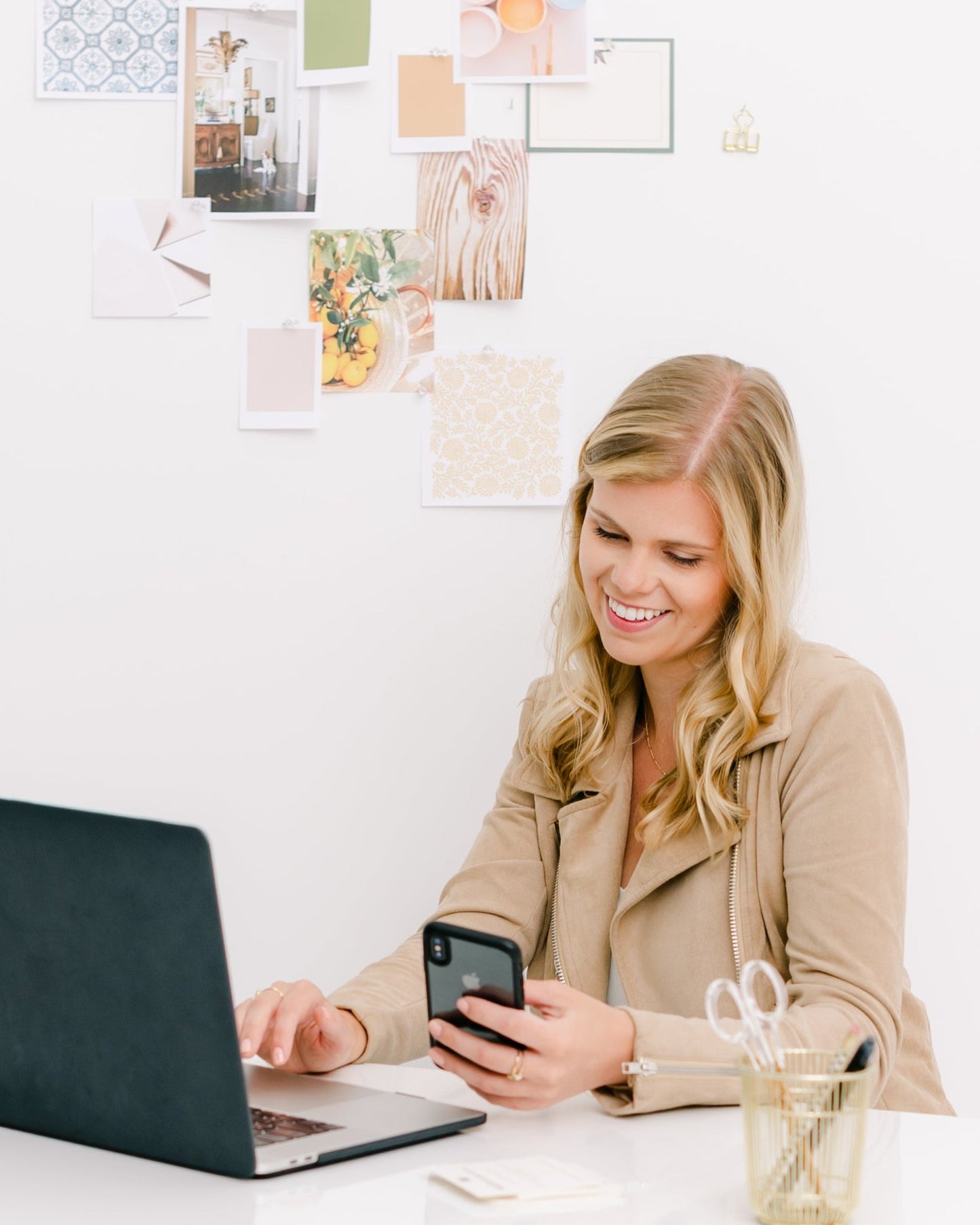 Woman using a laptop and phone at a desk with a white wall and decorative items in the background