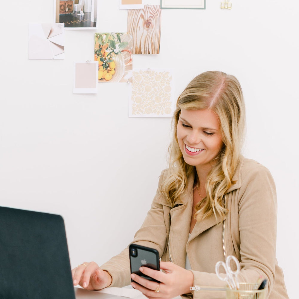 Woman using a laptop and phone at a desk with a white wall and decorative items in the background