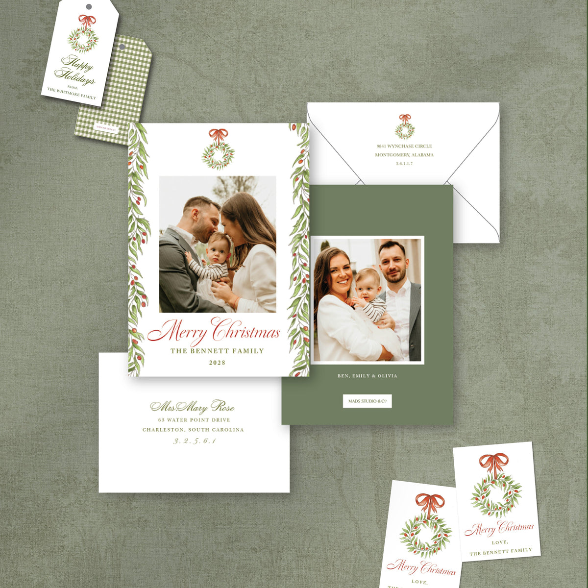 A coordinated Christmas stationery suite laid out on a textured gray-green background, including a main photo card with a wreath border and family photo, an envelope, a separate card, a small hang tag, and two smaller cards. The design uses white, muted sage green, red script text, and green/red holiday accents.