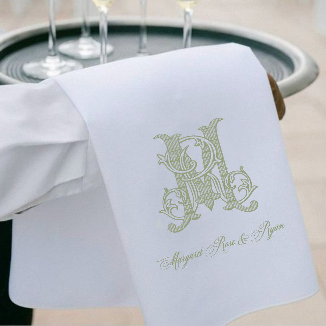 A white personalized tea towel with a green monogram and the names 'Margaret Rose & Ryan' printed on it, held by a person in a white shirt. In the background, a silver tray holds four filled champagne flutes.