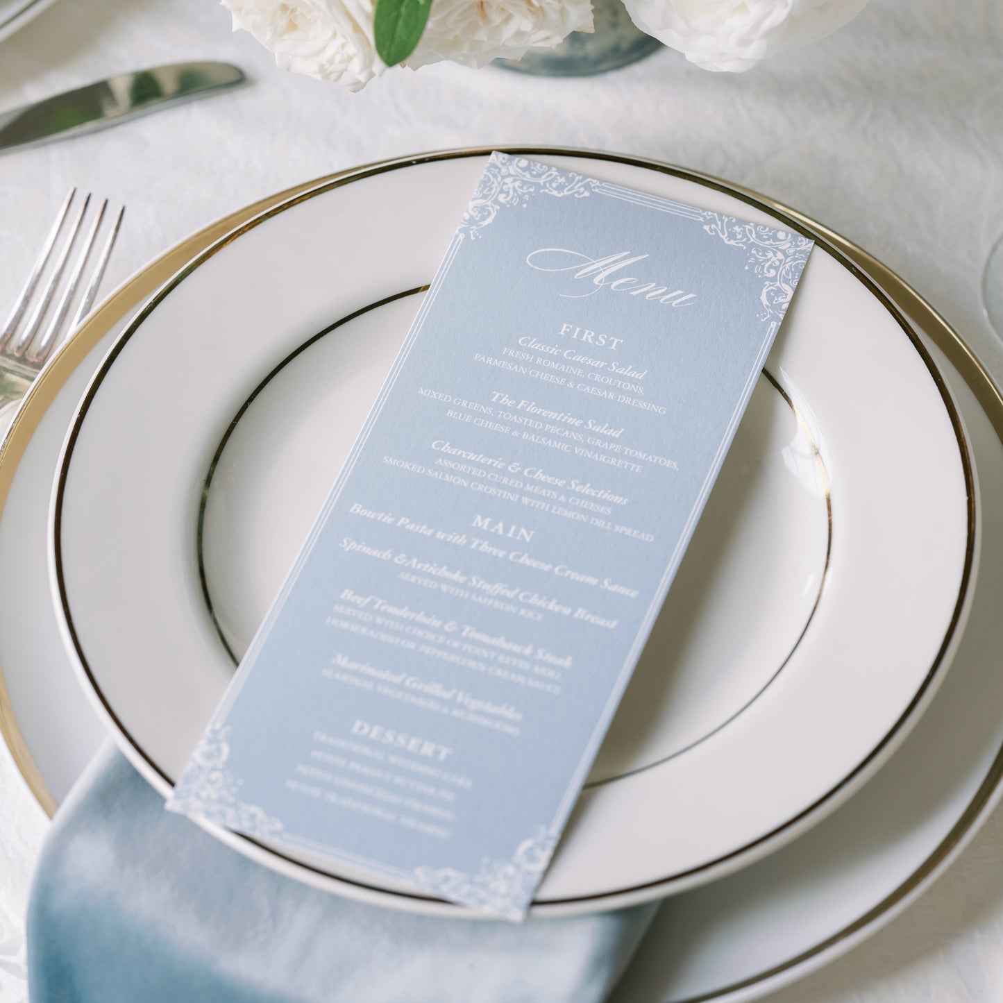 Table setting with white flowers, a blue menu card, and silverware on a white tablecloth.