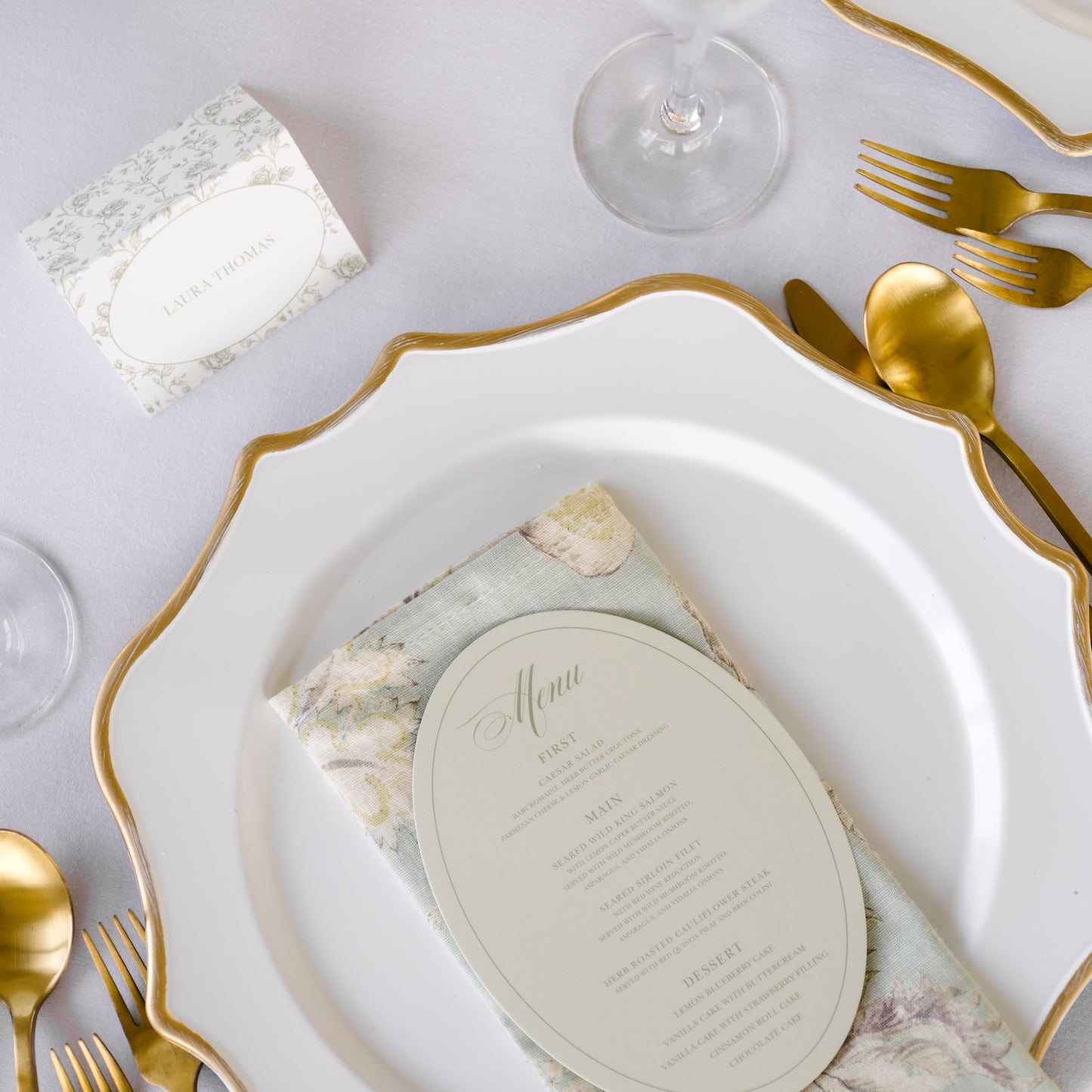 A styled place setting on a white tablecloth featuring a white plate with a gold scalloped rim, gold flatware, a floral-patterned napkin, and a round menu card.