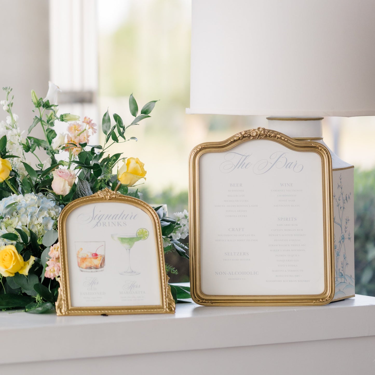A wedding stationery display featuring a gold-framed vertical guest book and a smaller matching gold-framed name card, styled next to a floral arrangement on a white console table.