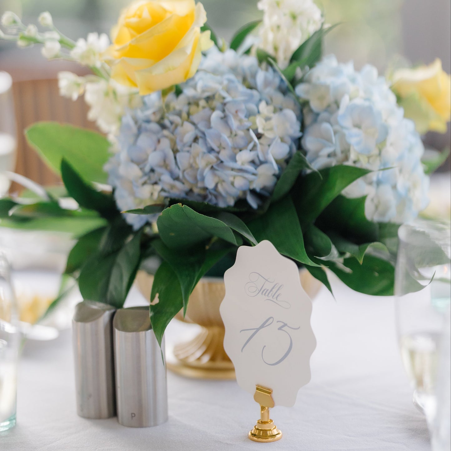 A floral table centerpiece with light blue hydrangeas, a yellow rose, white filler flowers, green leaves, and a small gold stand holding a paper table number card on a white tablecloth.